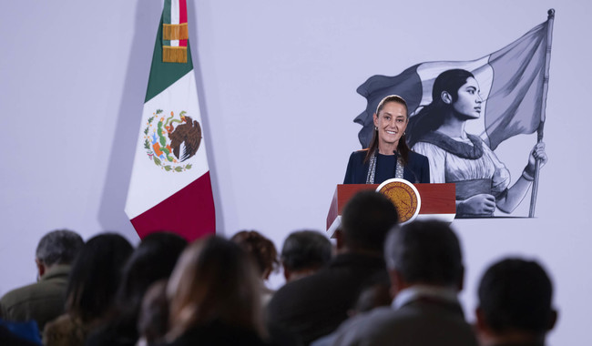 Claudia Sheinbaum en conferencia de prensa en Palacio Nacional, con bandera de México y mural de mujer indígena de fondo.